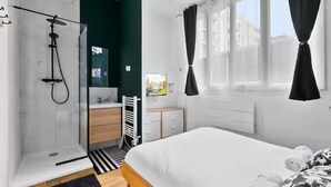 A modern hotel room with a shower, a bed, a vanity with a mirror, and a window with curtains.