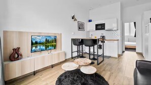 A modern living room with a flat-screen TV, a wooden cabinet, a black furry rug, and a dining area with a kitchen in the background.