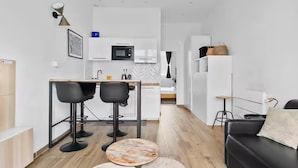 A modern kitchen with white cabinets, a microwave, and a sink. There are two black bar stools and a round table. A black sofa and a small round table are in the living area.
