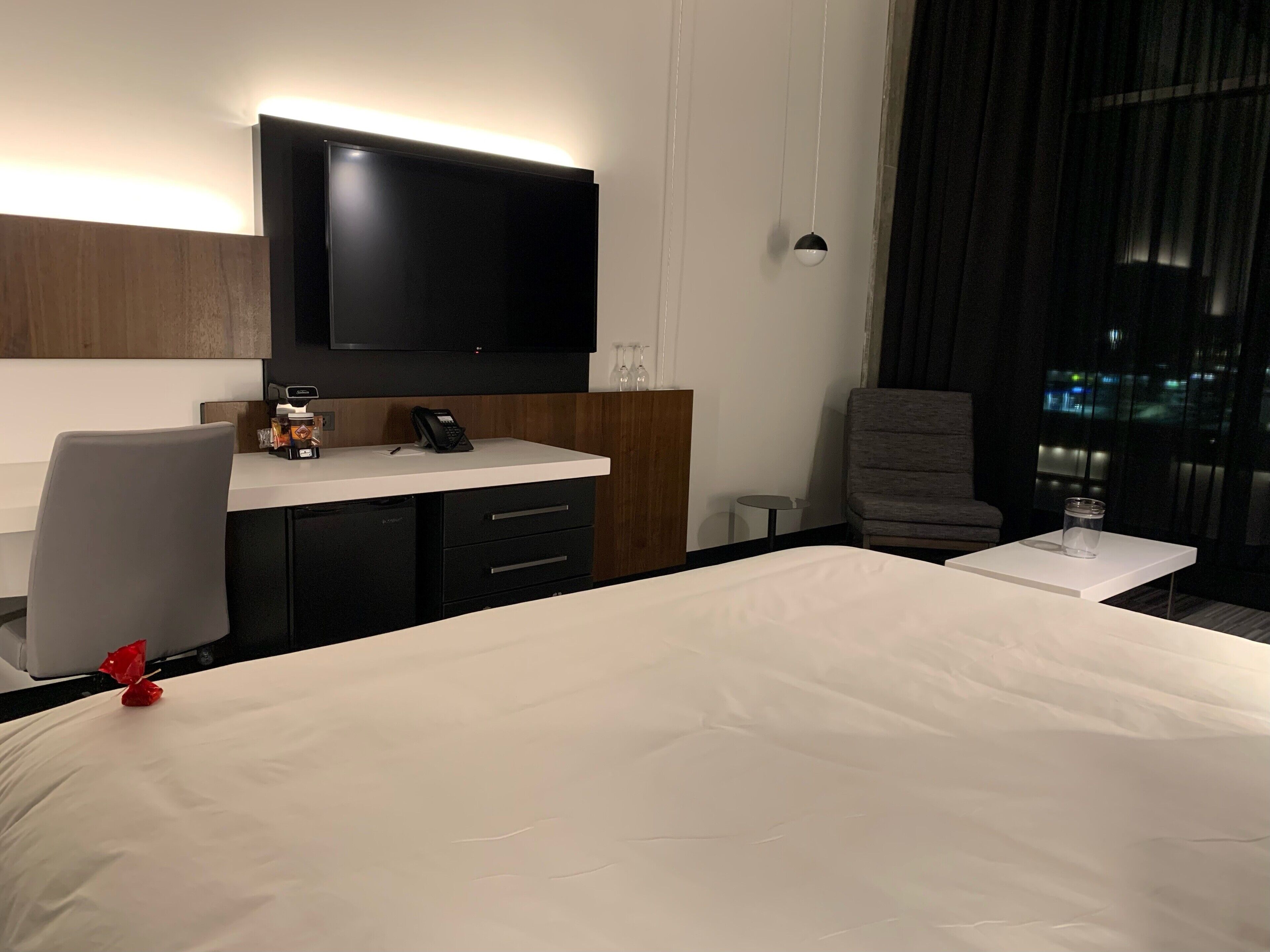 Et hotelværelse med en stor seng, et skrivebord med telefon, et fladskærms-tv monteret på væggen og et vindue med gardiner.