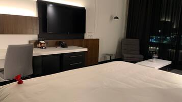 Et hotelværelse med en stor seng, et skrivebord med telefon, et fladskærms-tv monteret på væggen og et vindue med gardiner.