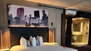 Quarto de hotel moderno com uma cama grande, obra de arte com vista da cidade na parede e banheiro visível.