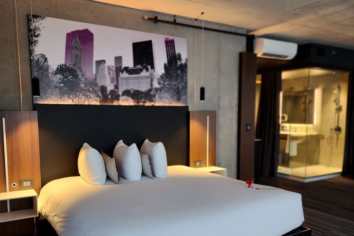 A modern hotel room with a large bed, cityscape artwork on the wall, and a visible bathroom.
