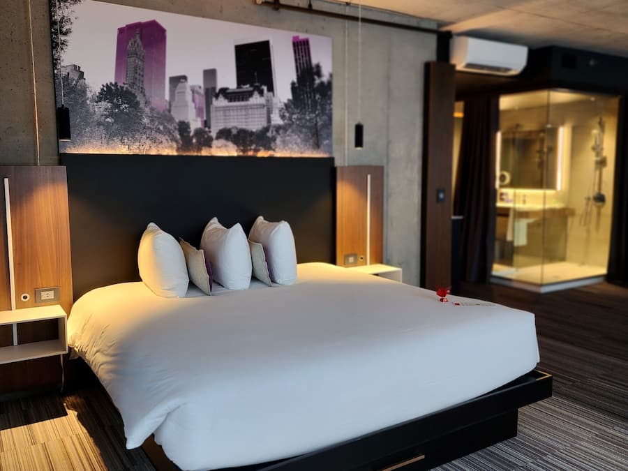 Une chambre d’hôtel moderne avec un grand lit, une œuvre d’art représentant un paysage urbain accrochée au mur et une salle de bain visible.