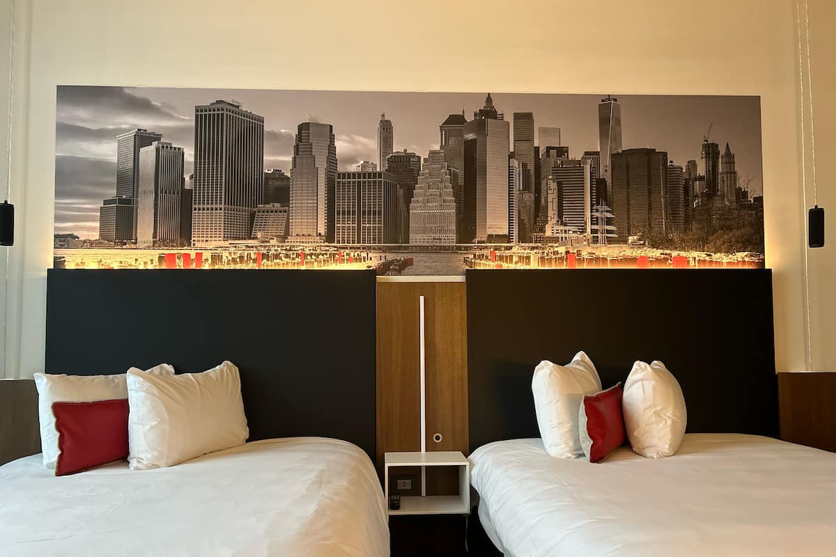 A hotel room with two beds, each with white bedding and red pillows, and a large cityscape mural on the wall.