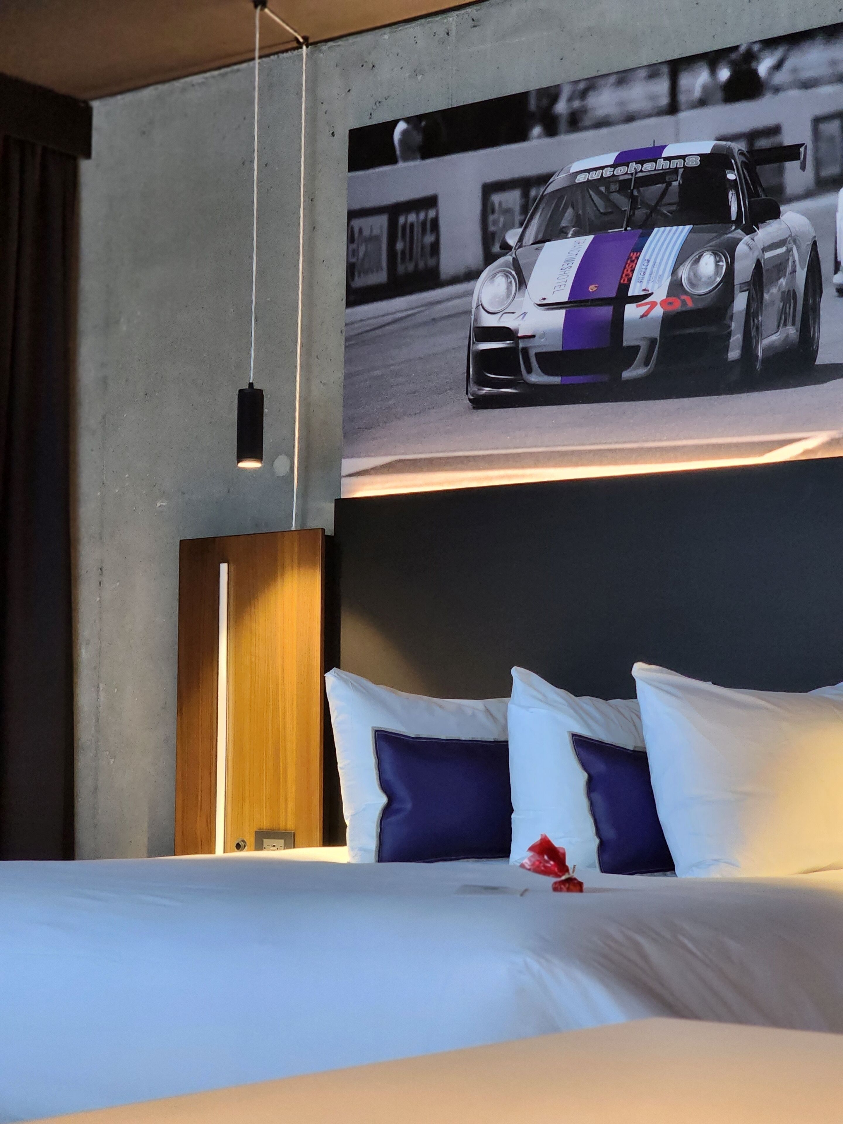 Une chambre d’hôtel avec un lit, des oreillers, une tête de lit et une affiche représentant une voiture de course accrochée au mur.