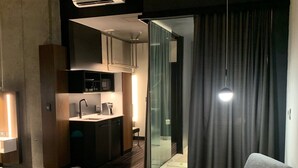 Quarto de hotel moderno com sofá, uma mesinha e um copo de água. Há uma área de cozinha com pia e armários, e uma porta coberta por cortinas que dá para outro cômodo.