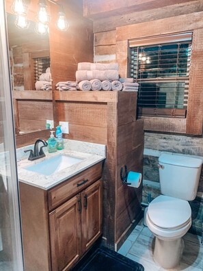 A bathroom with wooden walls, a sink with a mirror, a toilet, and a window with blinds. - Cabin at CM Cattleco (Newburg)