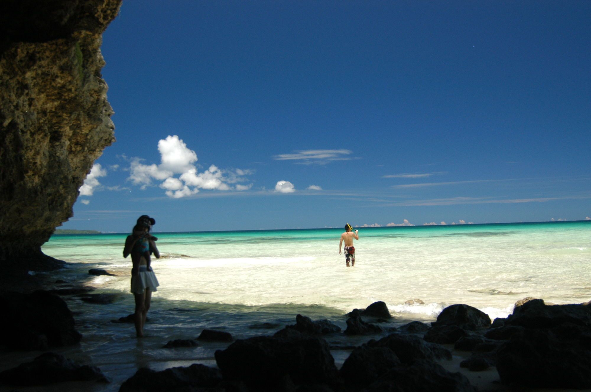 A beach scene with clear turquoise water, a rocky foreground, and a person standing in the distance.