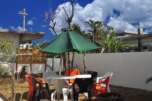 An outdoor seating area with a table and chairs, a green umbrella, and a white wall in the background. - Yukuiru (Miyakojima)