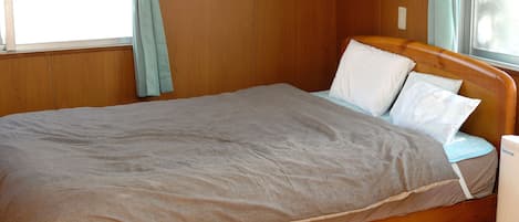 Una cama individual con estructura de madera, una colcha gris y almohadas blancas en una habitación con paredes de madera y un ventanal con cortinas.