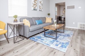 Una sala de estar con un sofá gris, una mesa de centro de madera, un tapete azul y una silla amarilla.