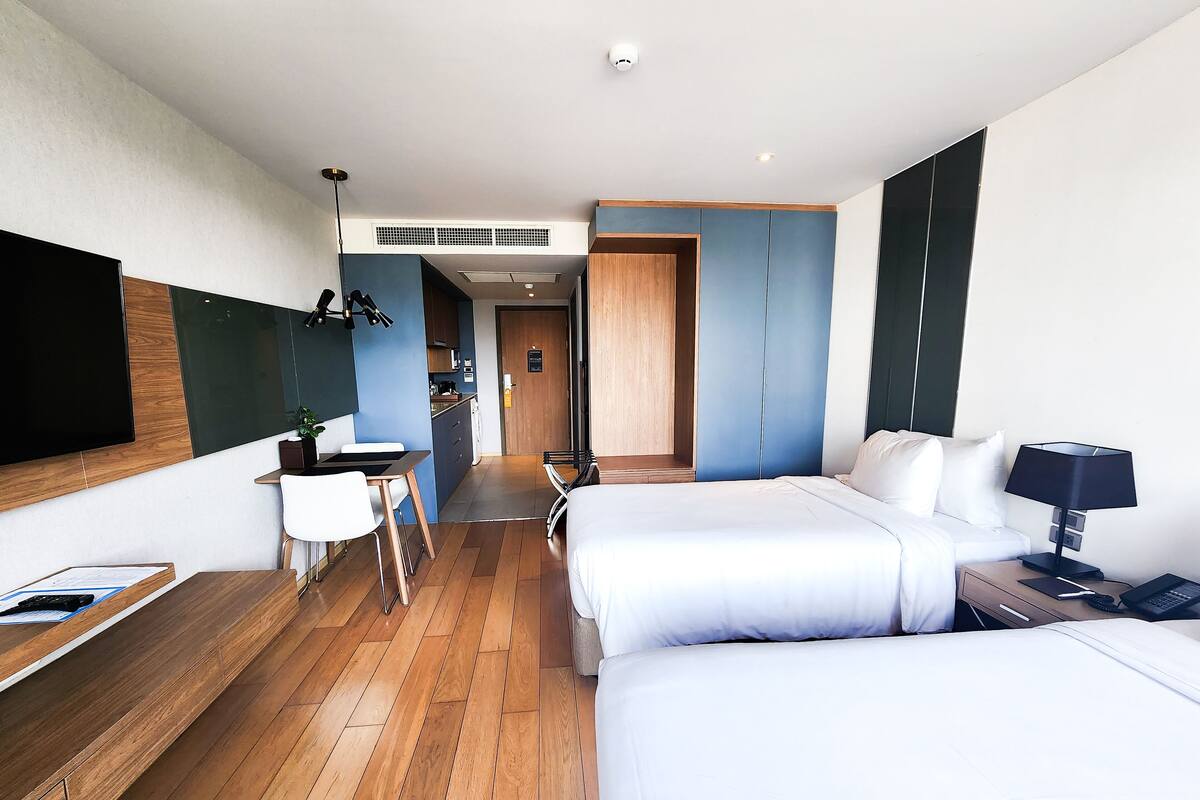 Une chambre d’hôtel moderne avec un plancher en bois, une grande télévision à écran plat, un bureau avec une chaise, un lit avec du linge de lit blanc et une table de chevet avec une lampe.