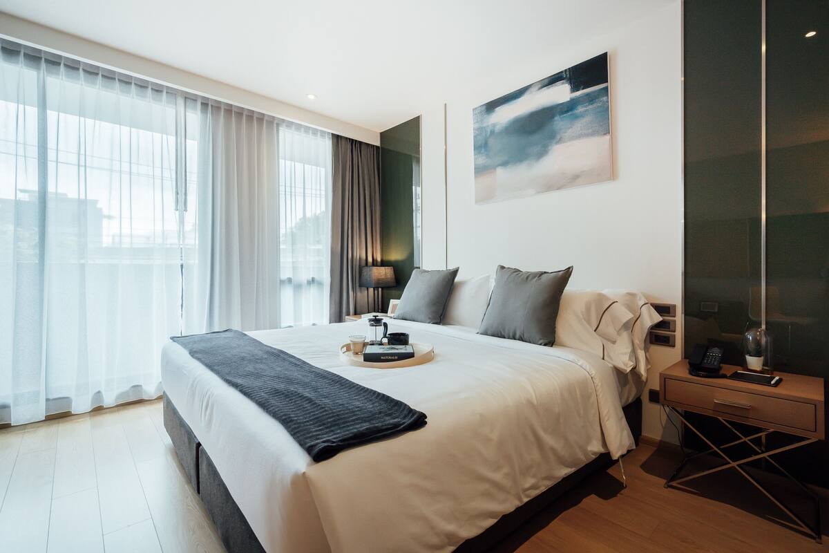 Une chambre d’hôtel moderne avec un grand lit, une table de chevet avec une lampe, une table basse en verre et un tableau au mur.