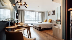 Ein modernes Hotelzimmer mit Essbereich, einem Bett mit weißer Daunendecke, einem Nachttisch mit Lampe und einem großen Fenster mit Vorhängen.