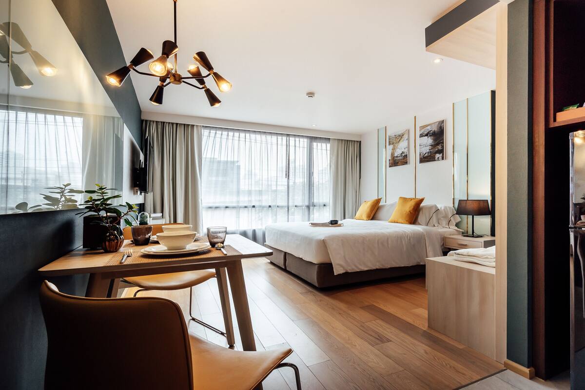 Une chambre d’hôtel moderne comprenant un coin repas, un lit avec une couette blanche, une table de chevet avec une lampe et une grande fenêtre avec des rideaux.