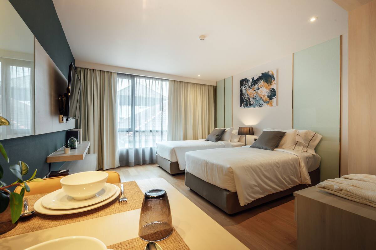 Une chambre d’hôtel moderne avec deux lits, un coin repas comprenant un saladier et des assiettes, et une grande fenêtre avec des rideaux.
