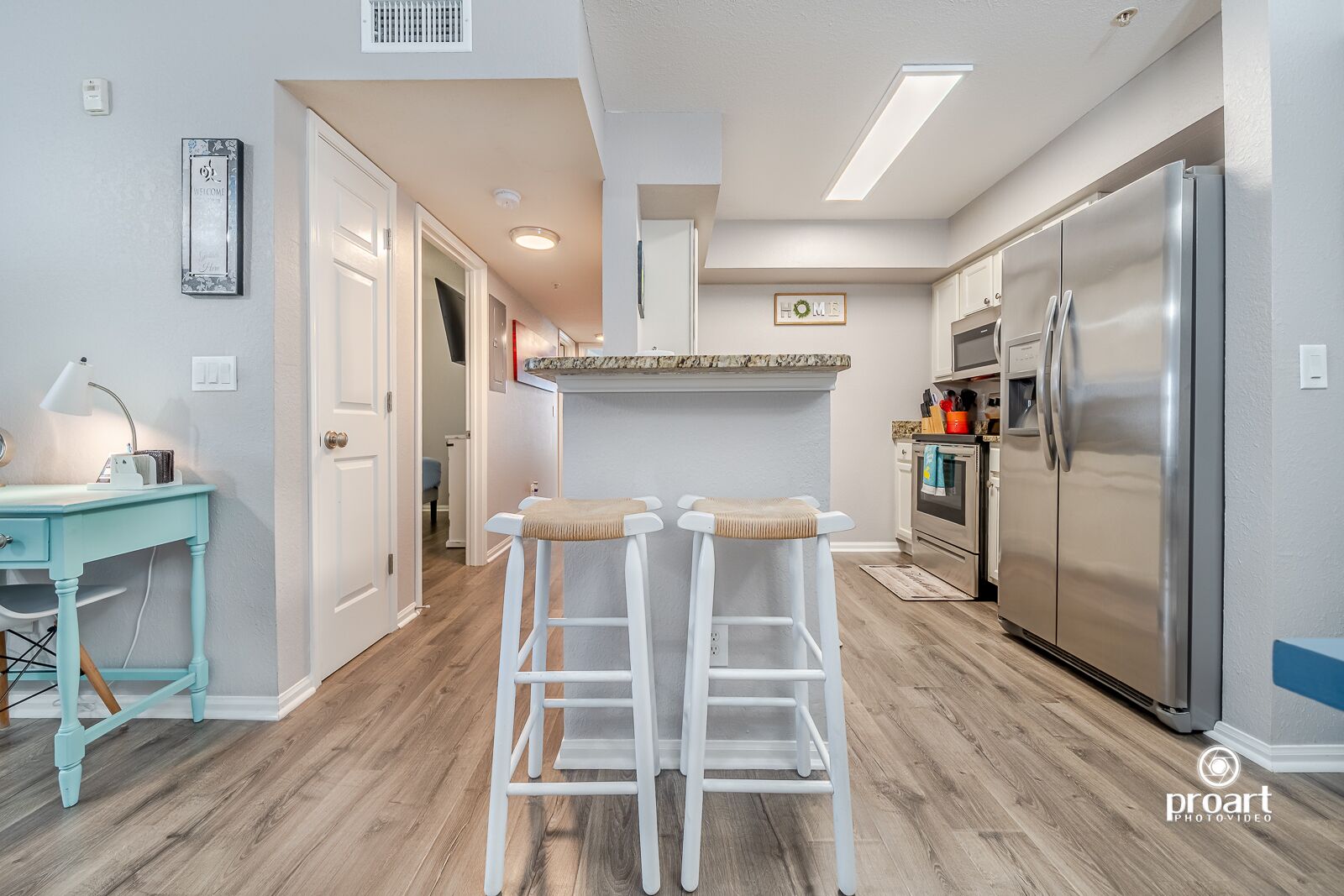 A modern kitchen with a stainless steel refrigerator, a built-in oven, and a granite countertop. There are two white bar stools and a teal desk with a lamp.