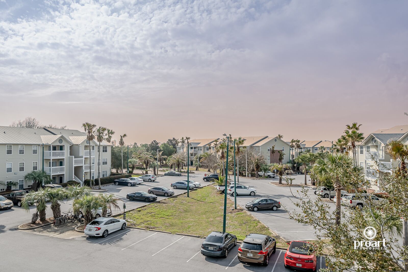 A parking lot with multiple cars and a row of apartment buildings with balconies and palm trees.