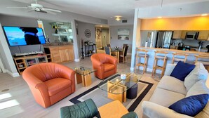 A modern living room with leather sofas, a glass coffee table, and a TV displaying a beach scene. - Pickleball, beach, pool, tennis, nextdoor to Publix, Junkanoo's and Fresh Catch. (Fort Myers Beach)