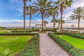 Stroll through the serene garden, lined with palm trees and neatly trimmed hedges, leading to a tranquil body of water and a picturesque gazebo.