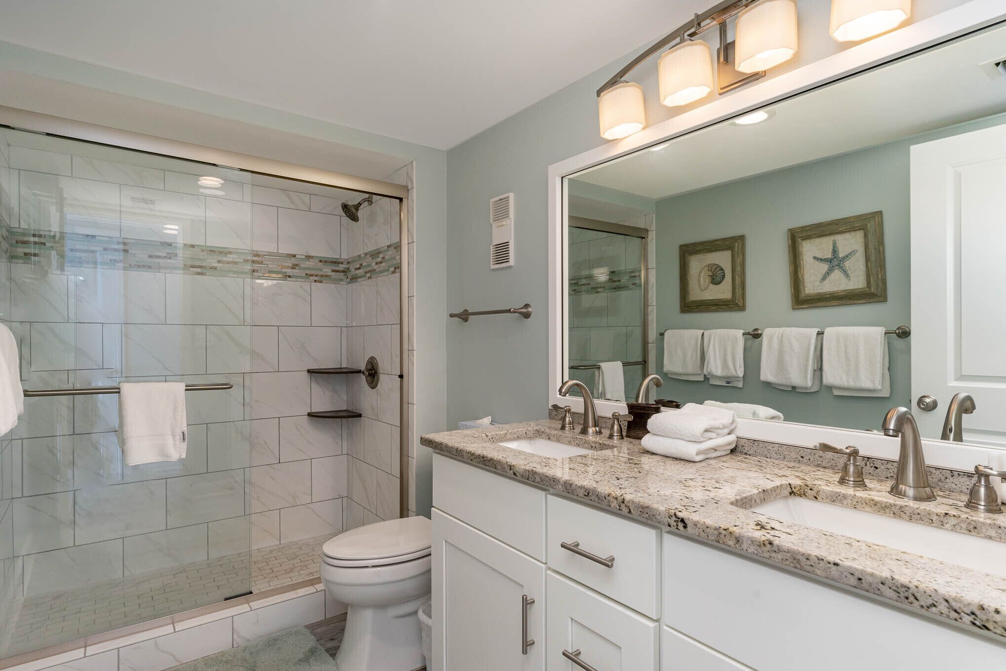 Een moderne badkamer met een glazen douchecabine, een wit toilet, een wastafel met granieten blad en een grote spiegel met decoratie van een zeester.