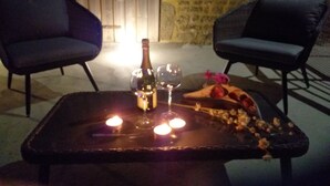 Una mesa con una botella, copas y un postre decorado con flores. Se observan dos sillas y un muro de piedra al fondo.