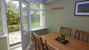 Ein Wintergarten mit Holztisch und -stühlen, einem gerahmten Bild an der Wand und Blick auf einen Garten durch Glastüren.