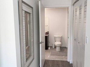 En smal gang der fører til et badeværelse med toilet, en lille hylde med håndklæder og et spejl.