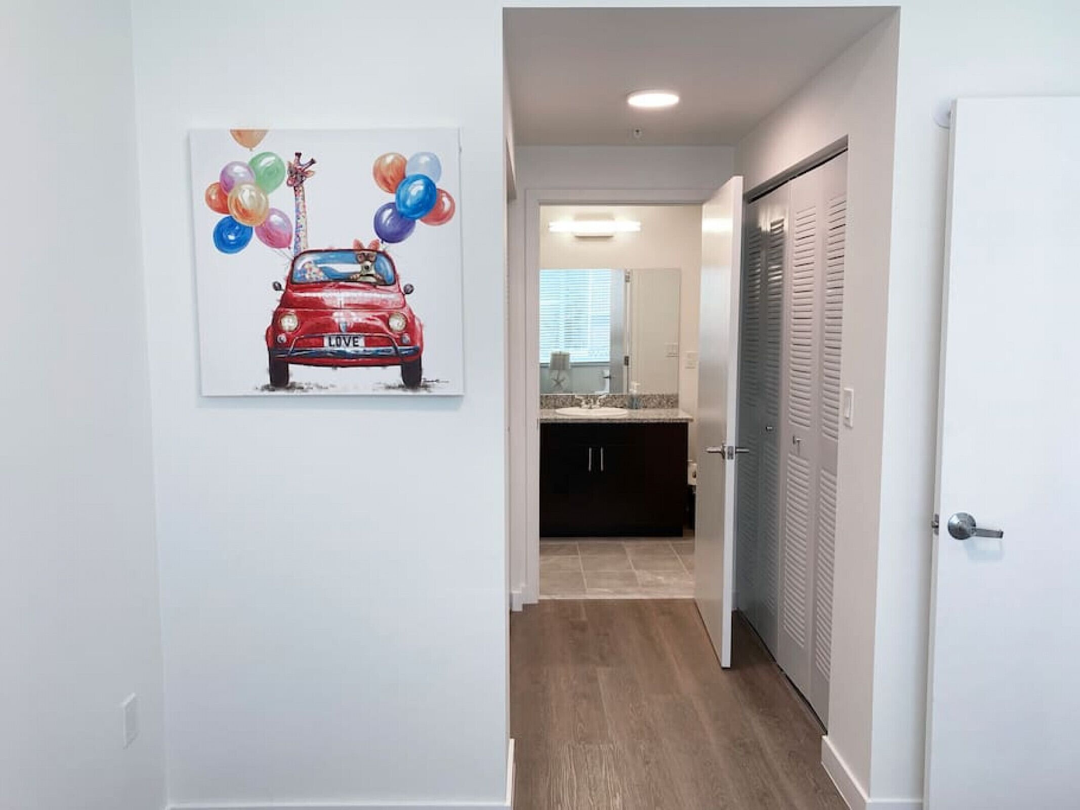 Un couloir orné d’un tableau représentant une voiture rouge et des ballons, une salle de bain avec un lavabo et un miroir, et une porte dotée de panneaux à persiennes.