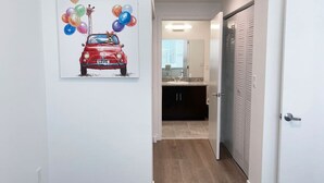 En hall med en målning av en röd bil och ballonger, ett badrum med handfat och spegel samt en dörr med lamellpaneler.