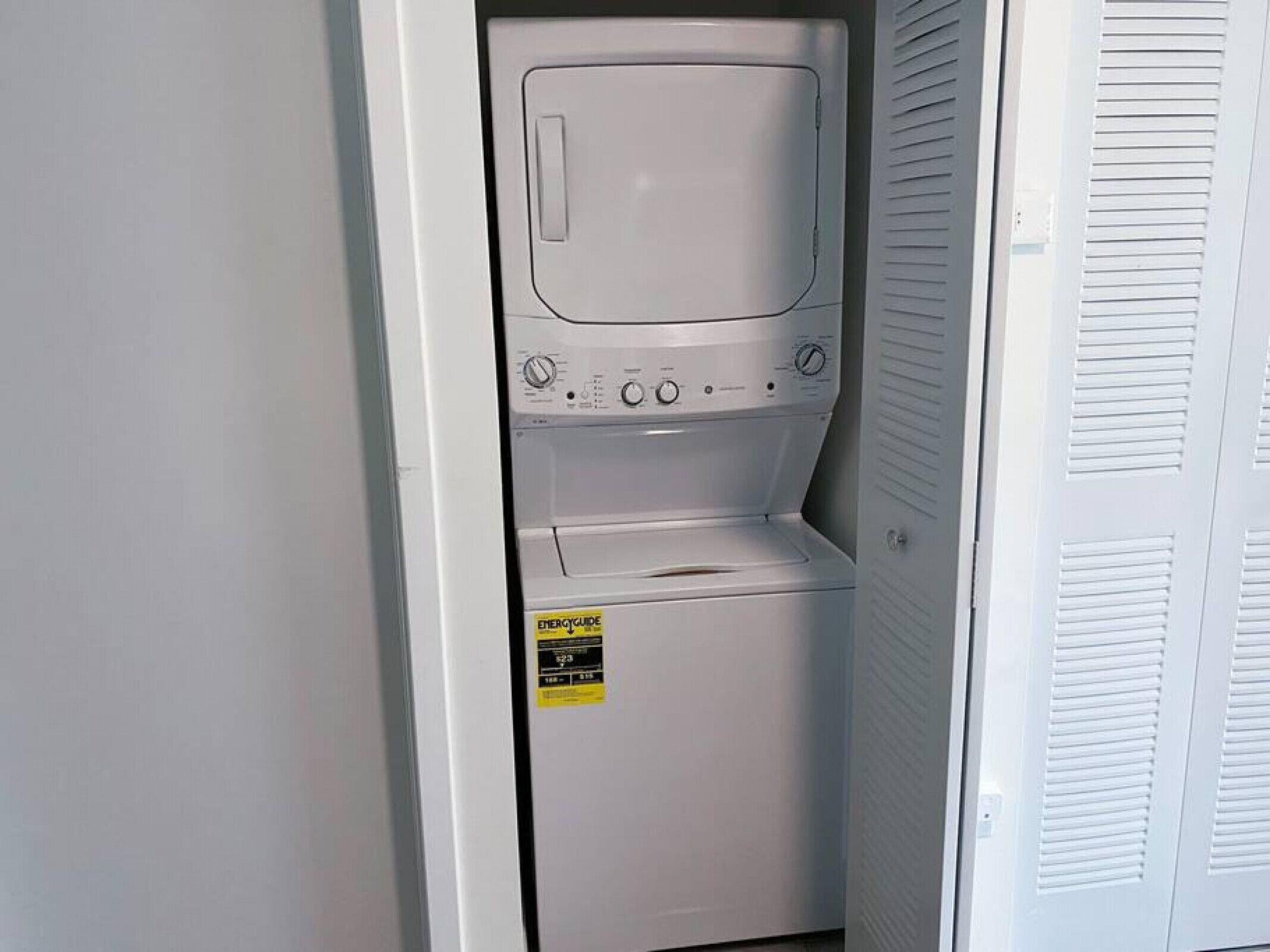 Une petite buanderie blanche, équipée d’un lave-linge et d’un sèche-linge à chargement frontal, d’une étiquette d’efficacité énergétique bien visible et d’une porte à panneaux ajourés.
