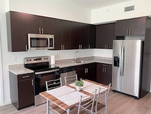 A modern kitchen with dark cabinets, stainless steel appliances, a granite countertop, and a dining area with a wooden table and chairs. - New, spacious and fully equipped 2bed/2bath apart (Miami)