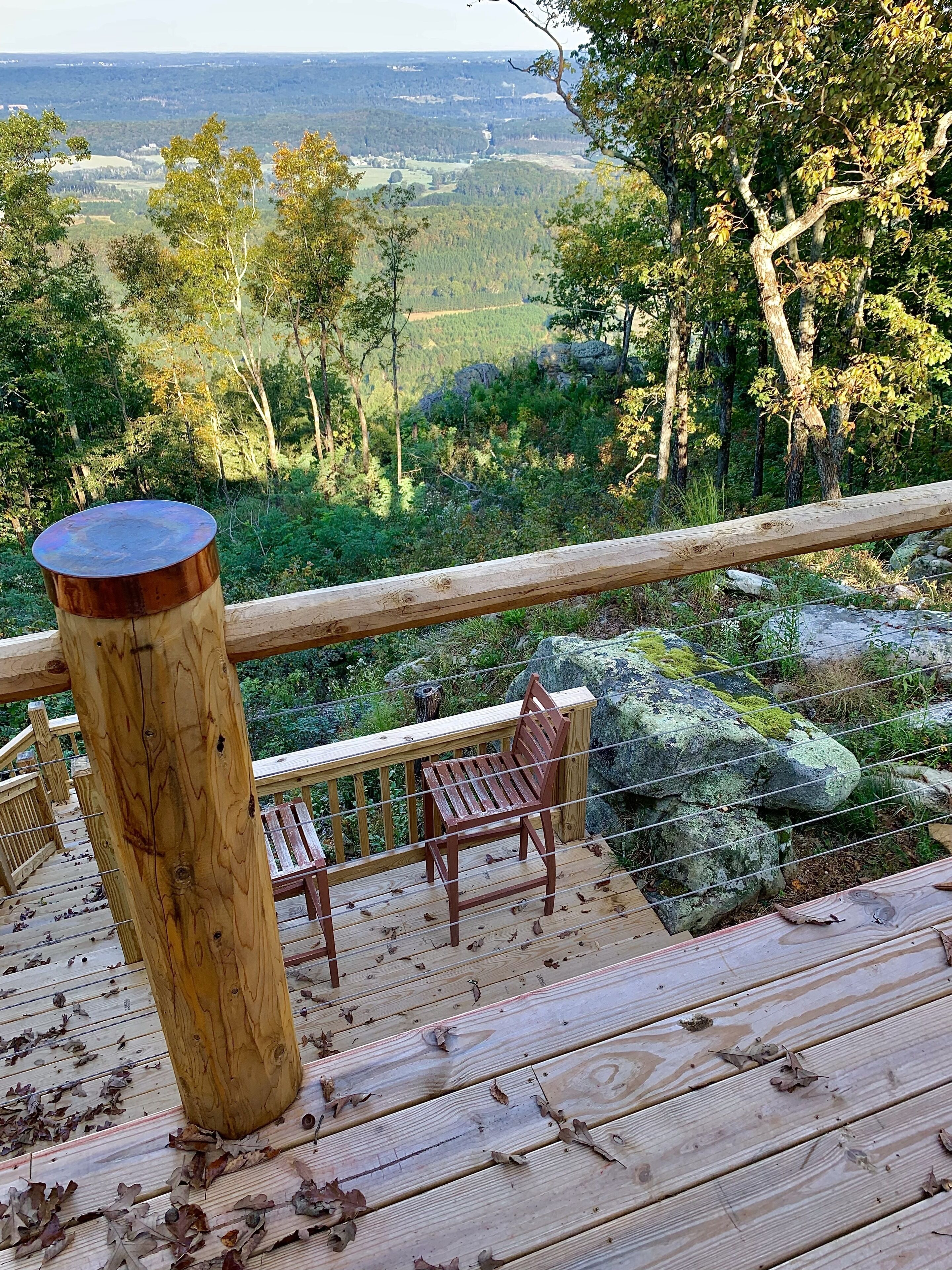 Una terraza de madera con un banco y una mesa, con vistas a un paisaje boscoso y colinas distantes.