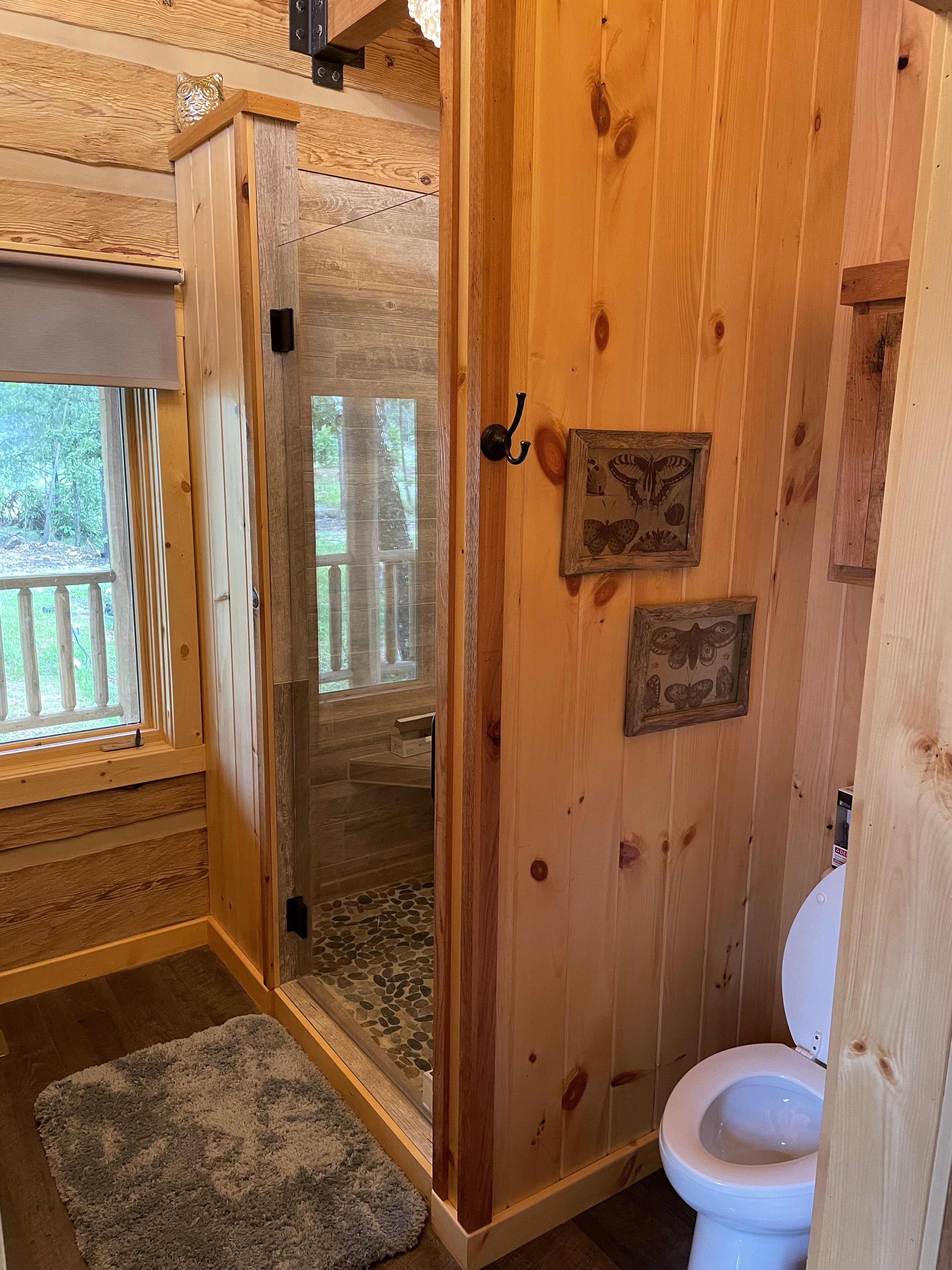 A wooden cabin bathroom with a glass shower, a toilet, and a window overlooking a deck.