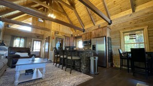 Interior amplio de cabaña con techo de madera, zona de comedor con encimera de piedra, cocina con electrodomésticos modernos y un rincón de estar acogedor.