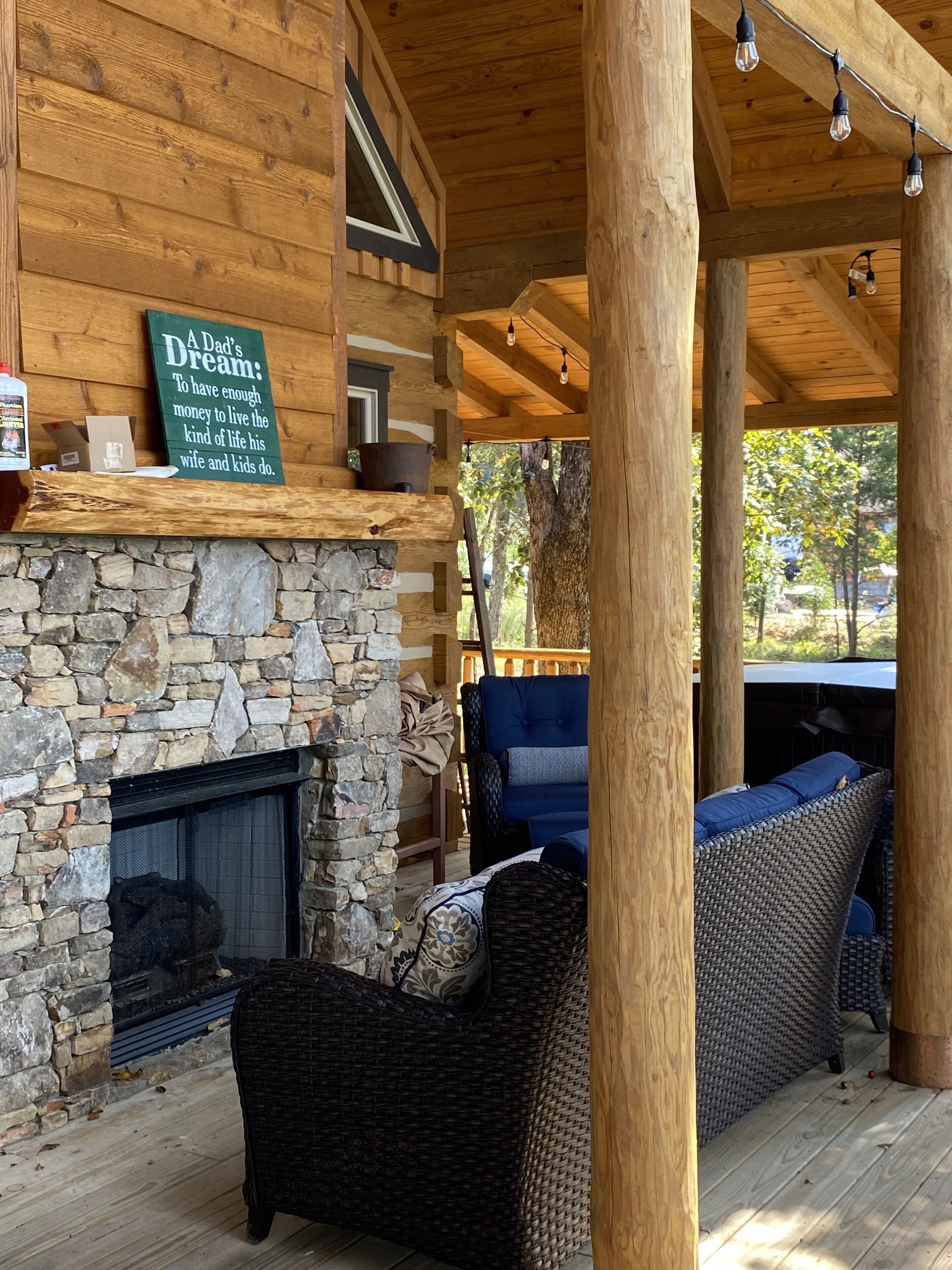 A cozy wooden cabin with a stone fireplace, wicker furniture, and a sign reading "A Dad's Dream: To have enough money to live the kind of life his wife and kids do."