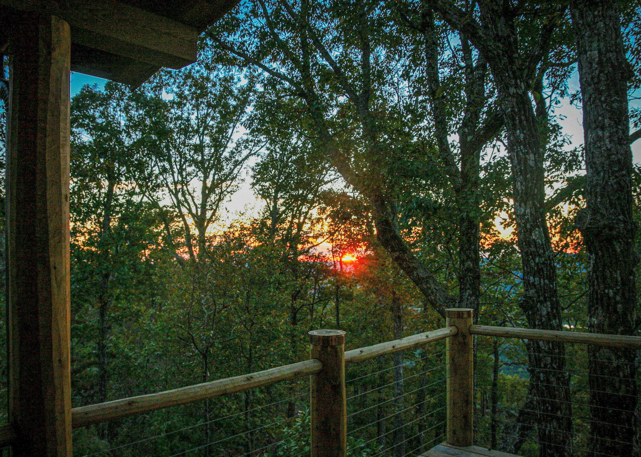 A wooden balcony railing overlooking a forested area with trees and a sunset in the background.
