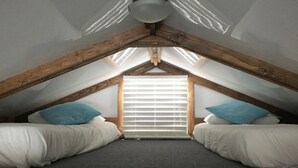 Ein kleines Schlafzimmer im Dachgeschoss mit schrägen Wänden, zwei Betten mit weißer Bettwäsche, einem Fenster mit Jalousien und einer Holzbalkenkonstruktion an der Decke.