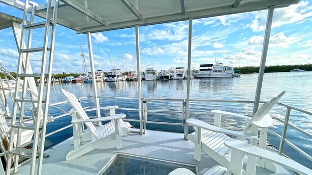 Eine überdachte Terrasse mit weißen Adirondack-Stühlen, einem Glasboden und Blick auf eine Marina mit Booten.
