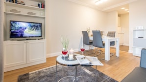 Een moderne woonkamer met een flatscreen-tv, een eethoek met een tafel en stoelen, en een boekenkast met boeken en decoratieve objecten.