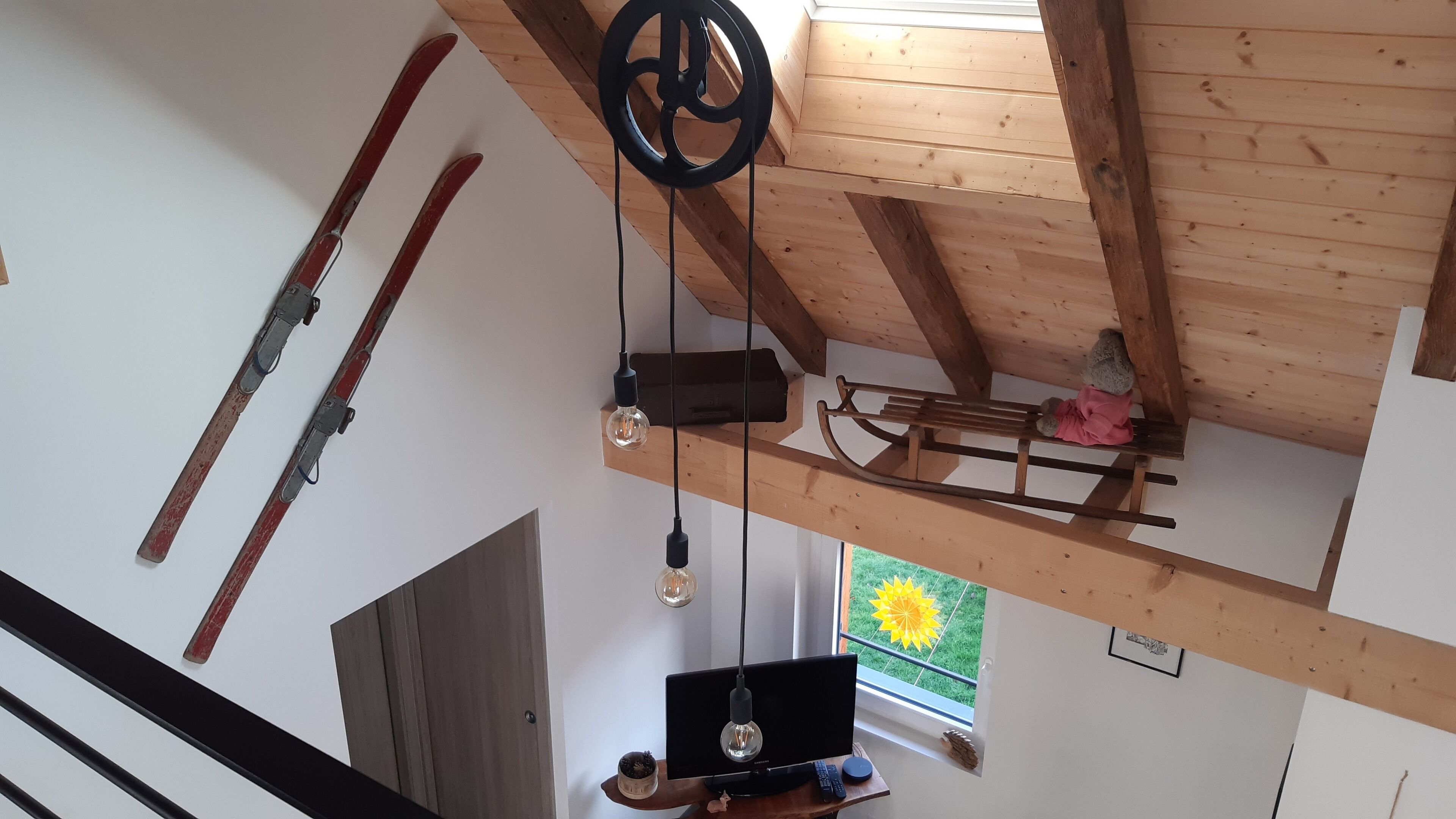 A room with a wooden ceiling, a TV on a wooden stand, and a window with a view of greenery.