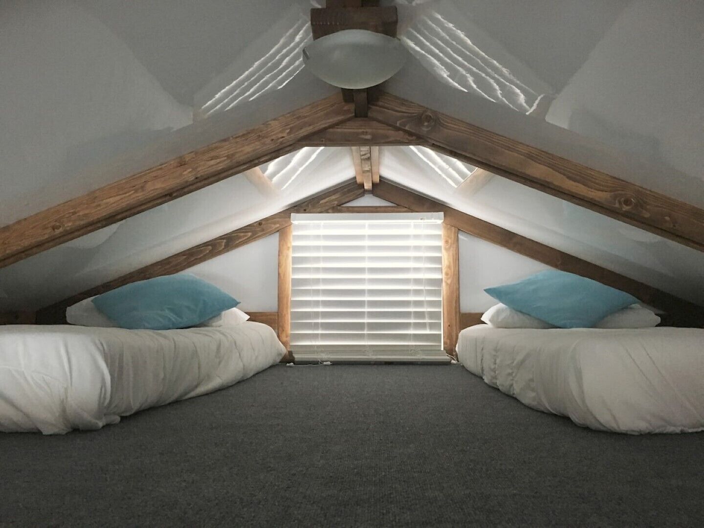 A small, attic-style bedroom with a sloped ceiling, two beds with white bedding, a window with blinds, and a wooden ceiling beam structure.