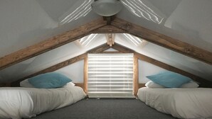A small, attic-style bedroom with a sloped ceiling, two beds with white bedding, a window with blinds, and a wooden ceiling beam structure.