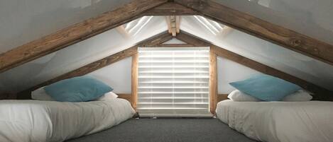A small, attic-style bedroom with a sloped ceiling, two beds with white bedding, a window with blinds, and a wooden ceiling beam structure.