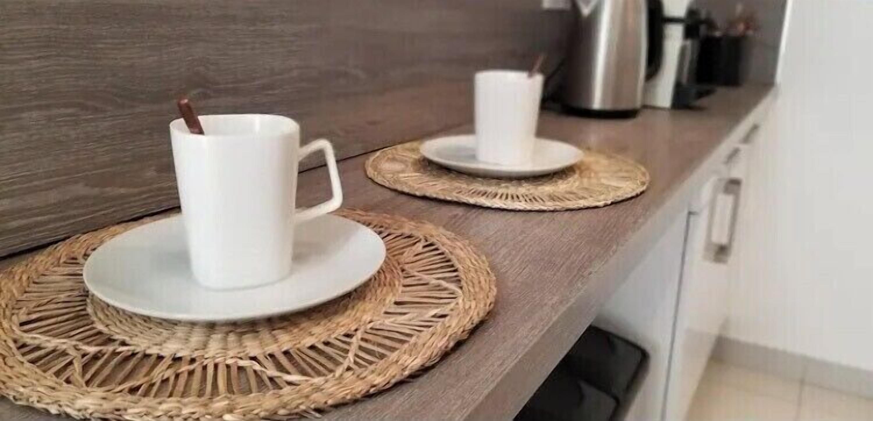 Two white cups on woven placemats on a kitchen counter.