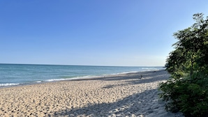 A wide sandy beach with gentle waves, a clear sky, and some greenery on the right.