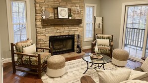 A cozy living room with a stone fireplace, wooden furniture, and large windows.