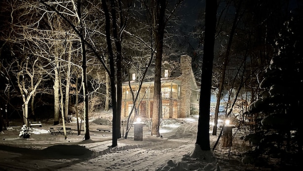 A snow-covered path leading to a stone house with a balcony, surrounded by bare trees.