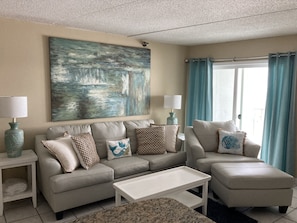 Relax in this stylish living room with a beige sofa, coffee table, and ottoman, complemented by a large abstract painting and a window with curtains.